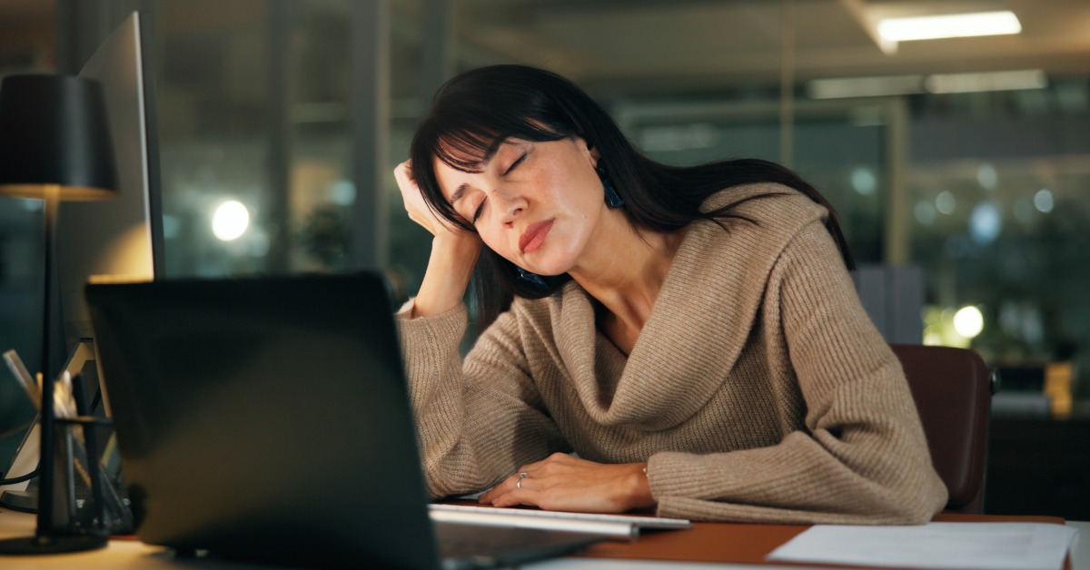 A young woman sits at her desk after hours as she rests her head in her hand. She's sleeping in front of her laptop.